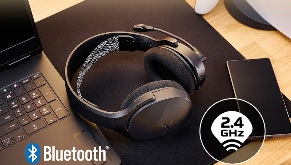 Na biurku znajdują się czarne słuchawki nauszne obok laptopa i smartfona. Widoczne są ikony Bluetooth i 2.4 GHz.