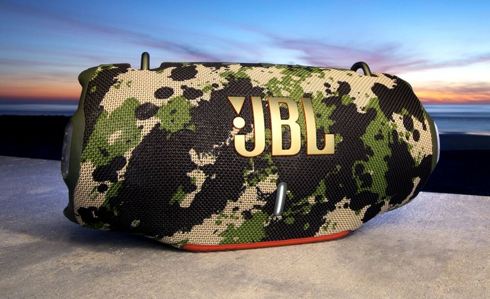 Głośnik mobilny z kamuflażowym wzorem i dużym logo JBL na siatce, ustawiony na tle zachodu słońca nad wodą.