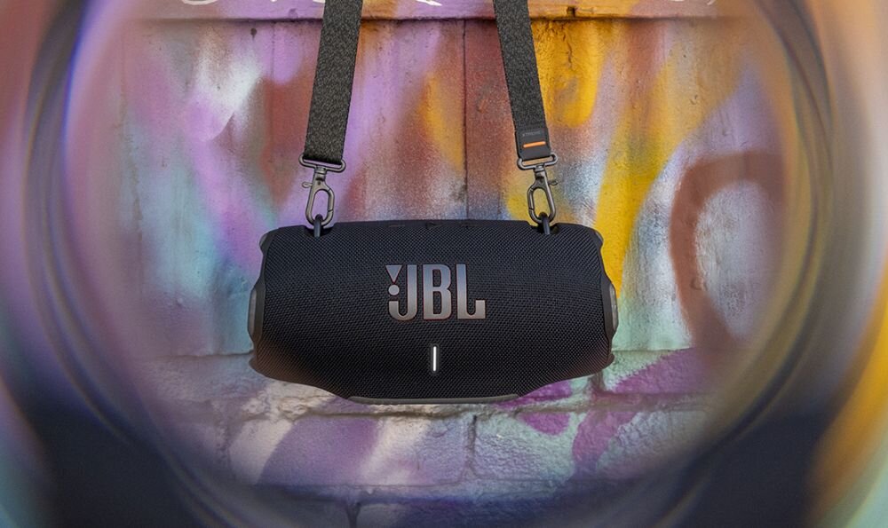 Czarny głośnik mobilny z logo JBL zawieszony na pasku, na tle kolorowej, graffiti ściany.