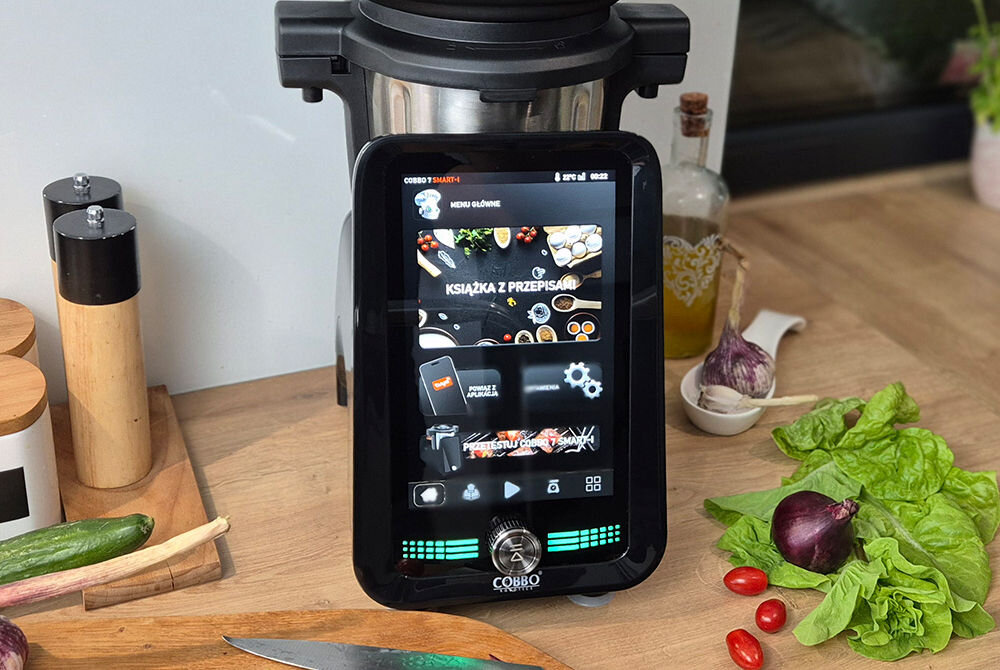 Na blacie kuchennym stoi robot wielofunkcyjny z dużym ekranem dotykowym, na którym widoczne jest menu z opcjami takimi jak \'Książka z przepisami\'. Obok urządzenia znajdują się warzywa, przyprawy i butelka z oliwą.