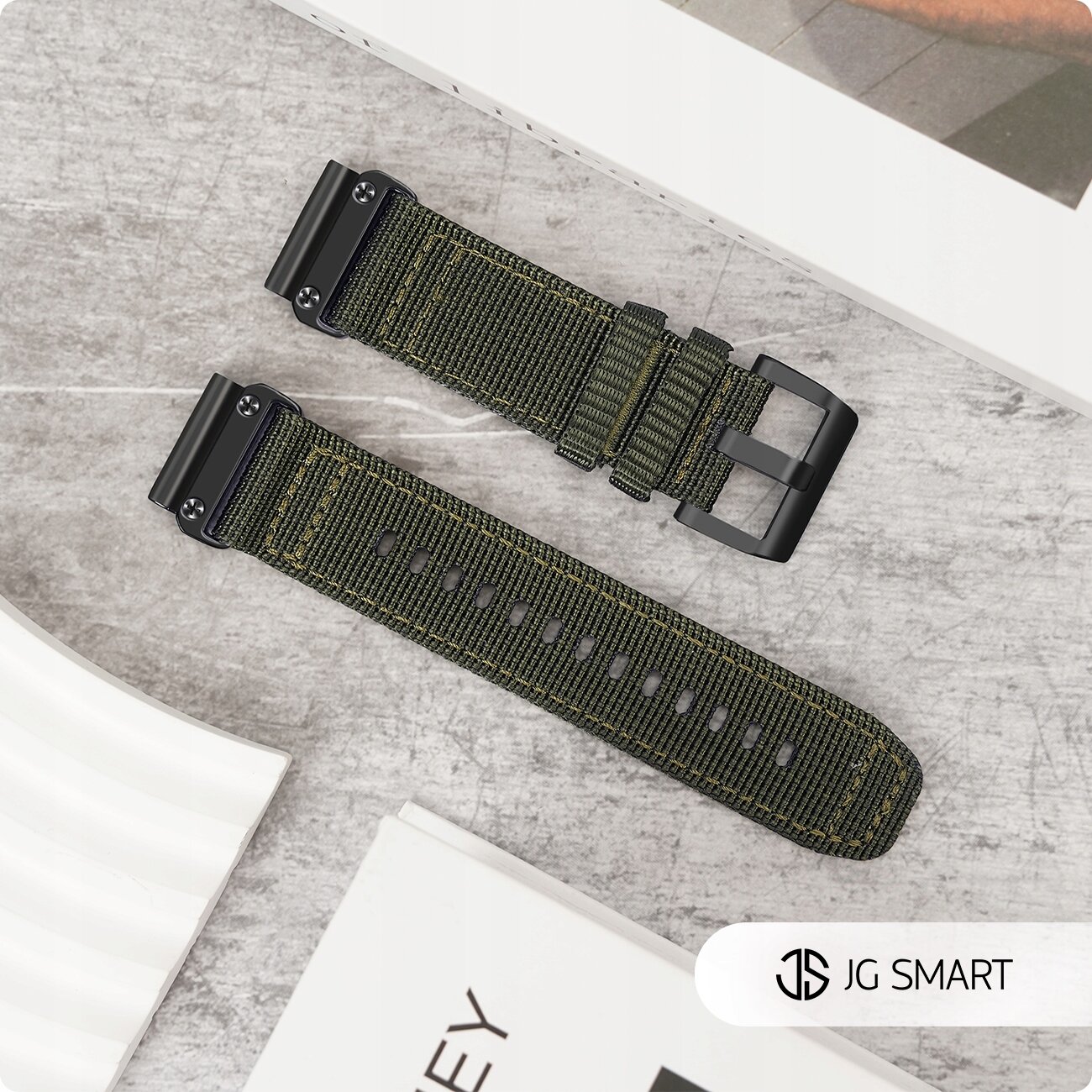 Na obrazie widoczny jest zielony pasek nylonowy z czarną klamrą, leżący na szarej powierzchni. W prawym dolnym rogu znajduje się logo JG SMART.