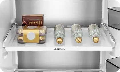 Wnętrze lodówki z półką oznaczoną jako \'Multi Tray\', na której znajdują się trzy butelki wody i opakowanie czekoladek. Półka jest wykonana z przezroczystego materiału z metalowymi uchwytami po bokach.
