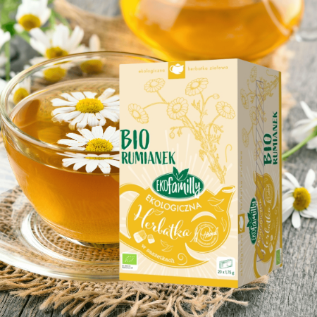 Na obrazie znajduje się opakowanie herbaty z napisem \'BIO RUMIANEK EKOFAMILY EKOLOGICZNA Herbatka\'. Obok opakowania stoi filiżanka z herbatą i kwiatami rumianku.