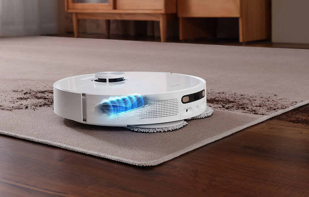 Na obrazie widoczny jest biały robot sprzątający na dywanie, który usuwa brud. W tle znajdują się drewniane meble.