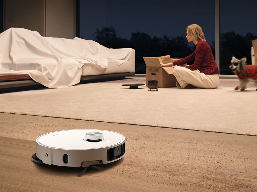Na podłodze w pomieszczeniu znajduje się biały robot sprzątający. W tle osoba siedzi na dywanie obok kartonu, a obok niej stoi pies ubrany w czerwony sweter.