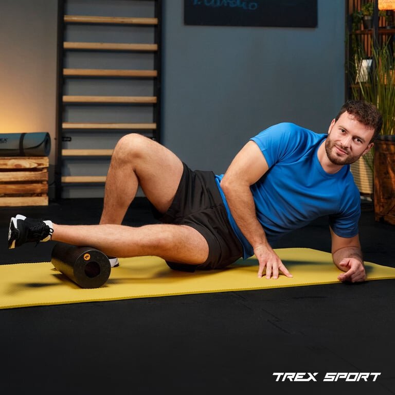 Osoba w niebieskiej koszulce leży na żółtej macie, używając czarnego wałka do masażu. W prawym dolnym rogu widoczne jest logo \'TREX SPORT\'.