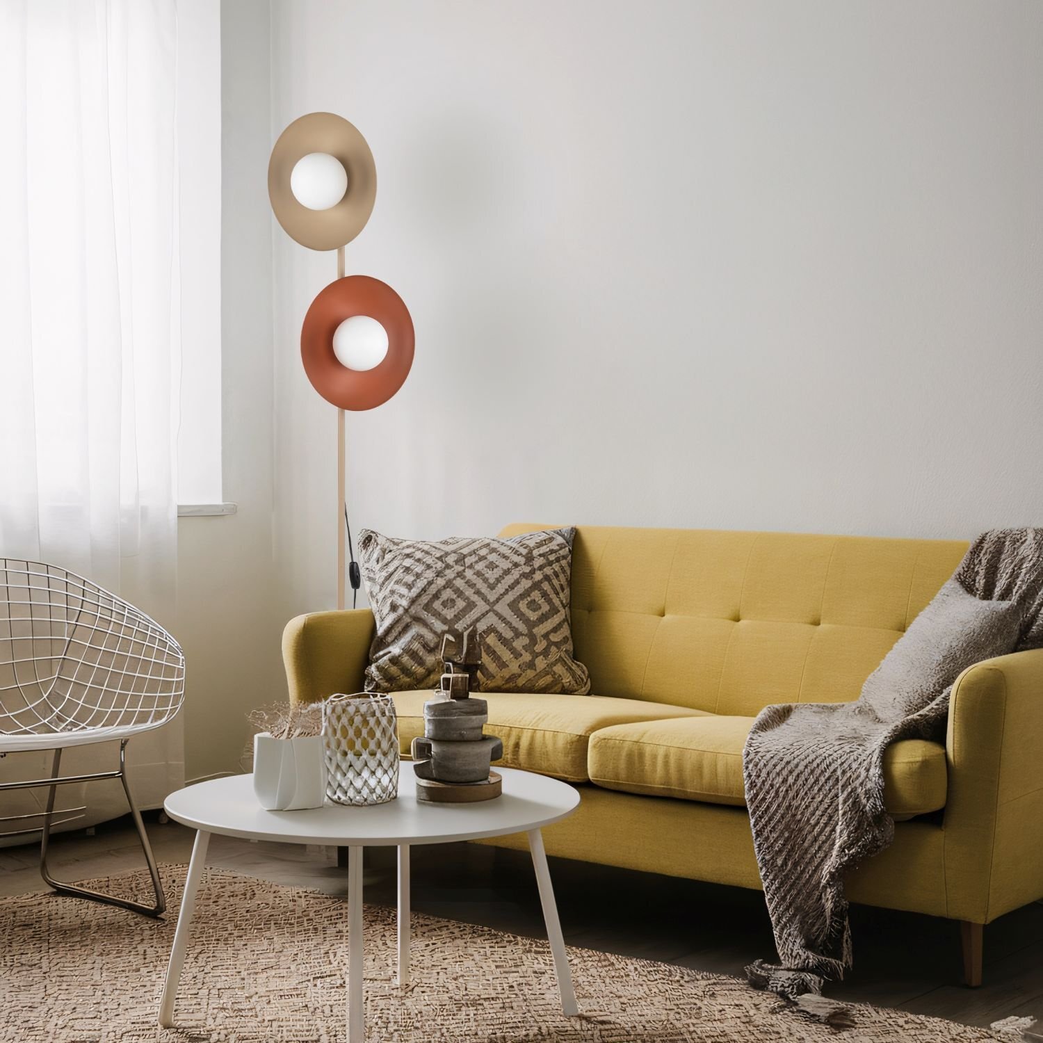 W salonie znajduje się żółta sofa z poduszką i kocem, obok której stoi nowoczesna lampa podłogowa z dwoma okrągłymi kloszami w kolorach beżowym i czerwonym. Przed sofą stoi biały stolik z dekoracjami, a w tle widać białą zasłonę.