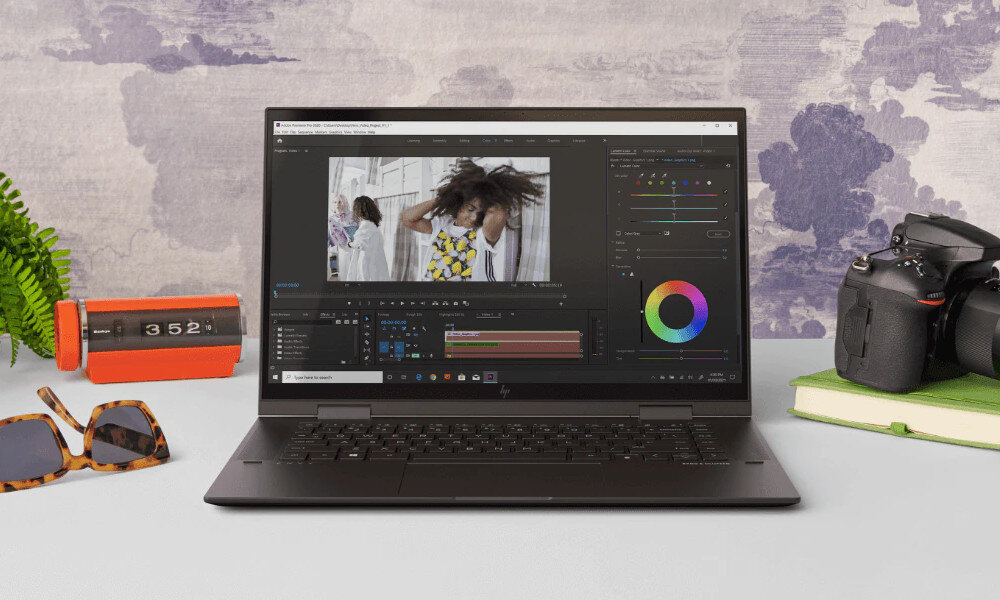 Na biurku znajduje się laptop z otwartym ekranem, na którym widoczny jest program do edycji wideo. Obok laptopa leżą okulary przeciwsłoneczne, aparat fotograficzny i zegar z wyświetlaczem 3:52.