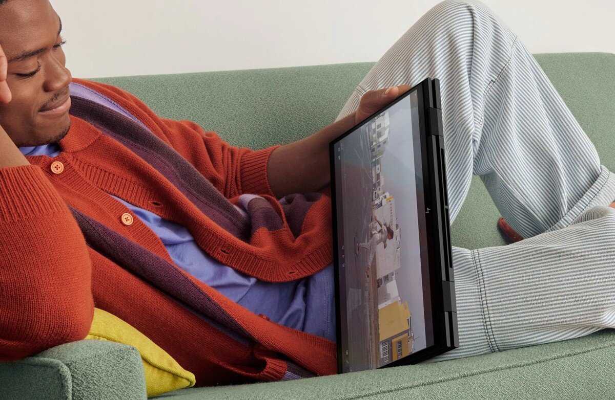 Osoba siedzi na kanapie, trzymając laptop w trybie tabletu, na ekranie widoczny jest film lub gra. Osoba ubrana jest w pomarańczowy sweter i pasiastą piżamę.
