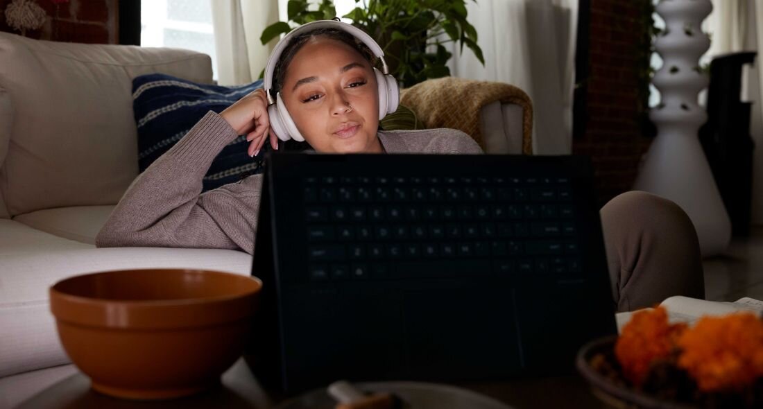 Osoba siedzi na kanapie, nosząc słuchawki, patrząc na ekran laptopa. W tle widać rośliny i zasłony, a na pierwszym planie znajduje się miska.