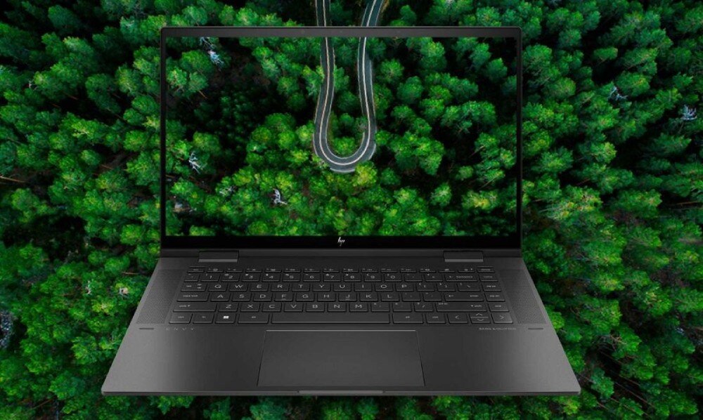 Laptop z otwartym ekranem, na którym wyświetlany jest obraz lasu z wijącą się drogą. Na obudowie widoczne jest logo producenta.