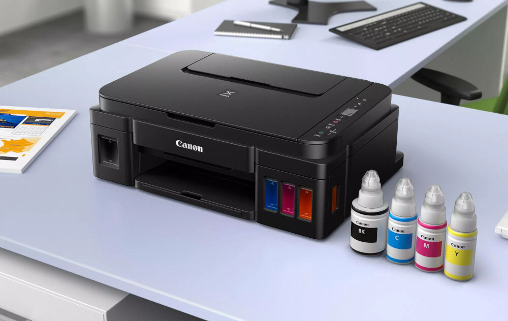 Urządzenie wielofunkcyjne CANON Pixma G2410 Druk w kolorze  prezentacja urządzenia na biurku w biurze obok butelki z atramentem ekonomiczny druk ilość stron na jednym zestawie zbiorników ułatwia zarządzanie kosztami swoboda drukowania