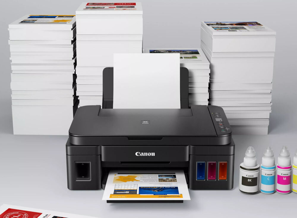 Urządzenie wielofunkcyjne CANON Pixma G2410 Druk w kolorze  prezentacja urządzenia na blacie za nim stos dokumentów obok butelki z atramentem prędkość druku kolor monochromatyczny odbitki wymiary pamiątkowe albumy