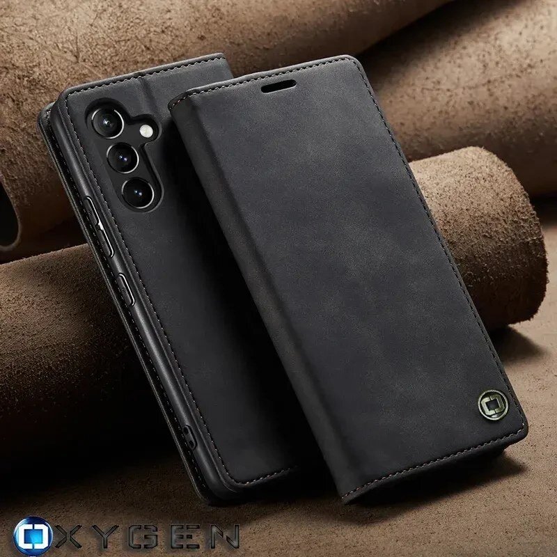 Na zdjęciu widoczne jest czarne etui na telefon z trzema wycięciami na aparaty oraz logo OXYGEN. Etui ma teksturę przypominającą skórę i jest ustawione na tle brązowych cylindrycznych elementów.