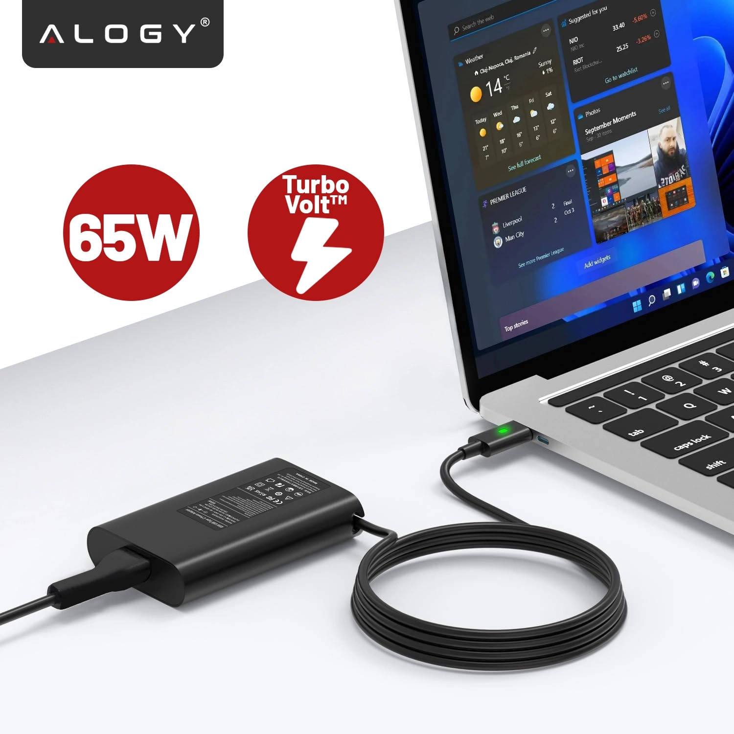 Na obrazie widoczna jest czarna ładowarka podłączona do laptopa z ekranem wyświetlającym interfejs systemu operacyjnego. Obok ładowarki znajdują się dwa czerwone symbole z napisami \'65W\' i \'Turbo Volt™\'.