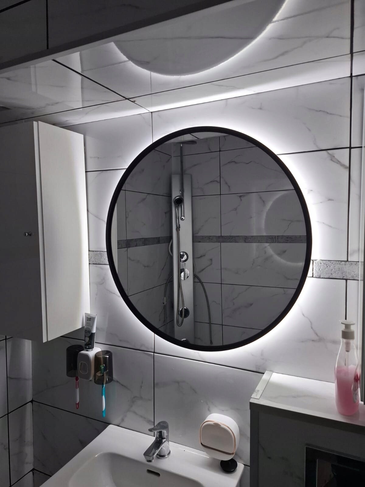Okrągłe lustro LED w czarnej aluminiowej ramie, zamontowane na ścianie wyłożonej płytkami imitującymi marmur, z widocznym odbiciem prysznica. Pod lustrem znajduje się umywalka z kranem oraz dozownikiem mydła.