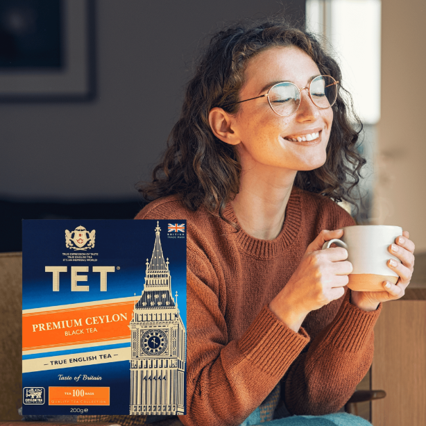 Kobieta w okularach trzyma filiżankę, obok niej opakowanie herbaty TET Premium Ceylon Black Tea z wizerunkiem Big Bena. Na opakowaniu widnieje napis: \'PREMIUM CEYLON BLACK TEA, TRUE ENGLISH TEA, Taste of Britain, 100 bags\'.