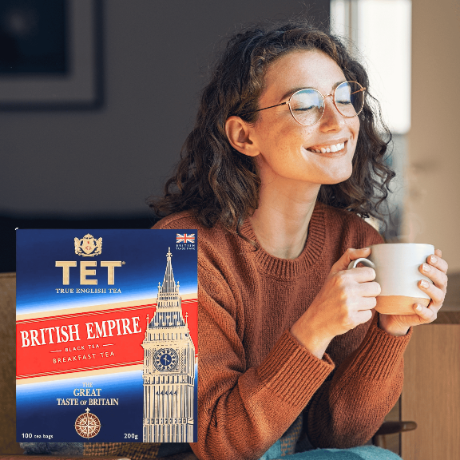 Kobieta w okularach trzyma kubek, obok niej opakowanie herbaty TET British Empire z wizerunkiem Big Bena. Na opakowaniu widnieje napis: \'TET True English Tea, British Empire, Black Tea, Breakfast Tea, Great Taste of Britain, 100 tea bags, 200g\'.