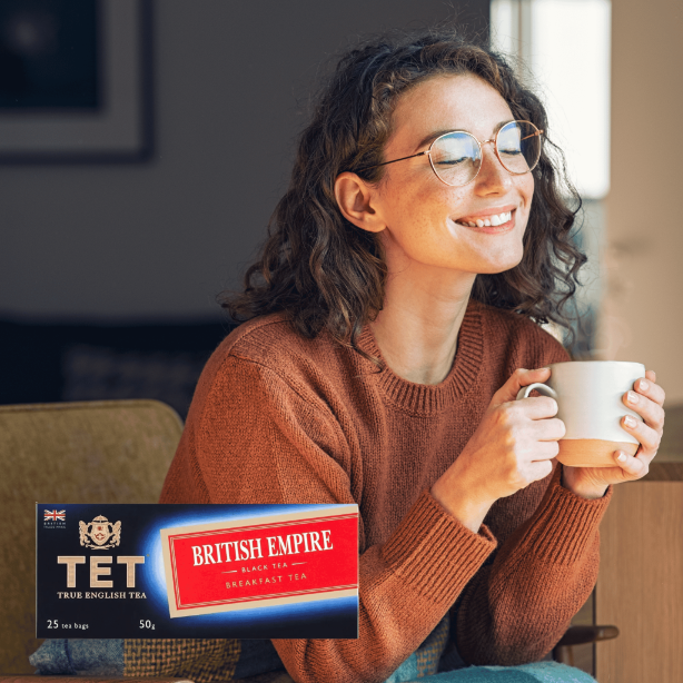 Osoba siedzi z filiżanką w dłoniach, uśmiechając się. Na pierwszym planie widoczna jest paczka herbaty z napisem \'TET TRUE ENGLISH TEA BRITISH EMPIRE BLACK TEA BREAKFAST TEA 25 tea bags 50g\'.