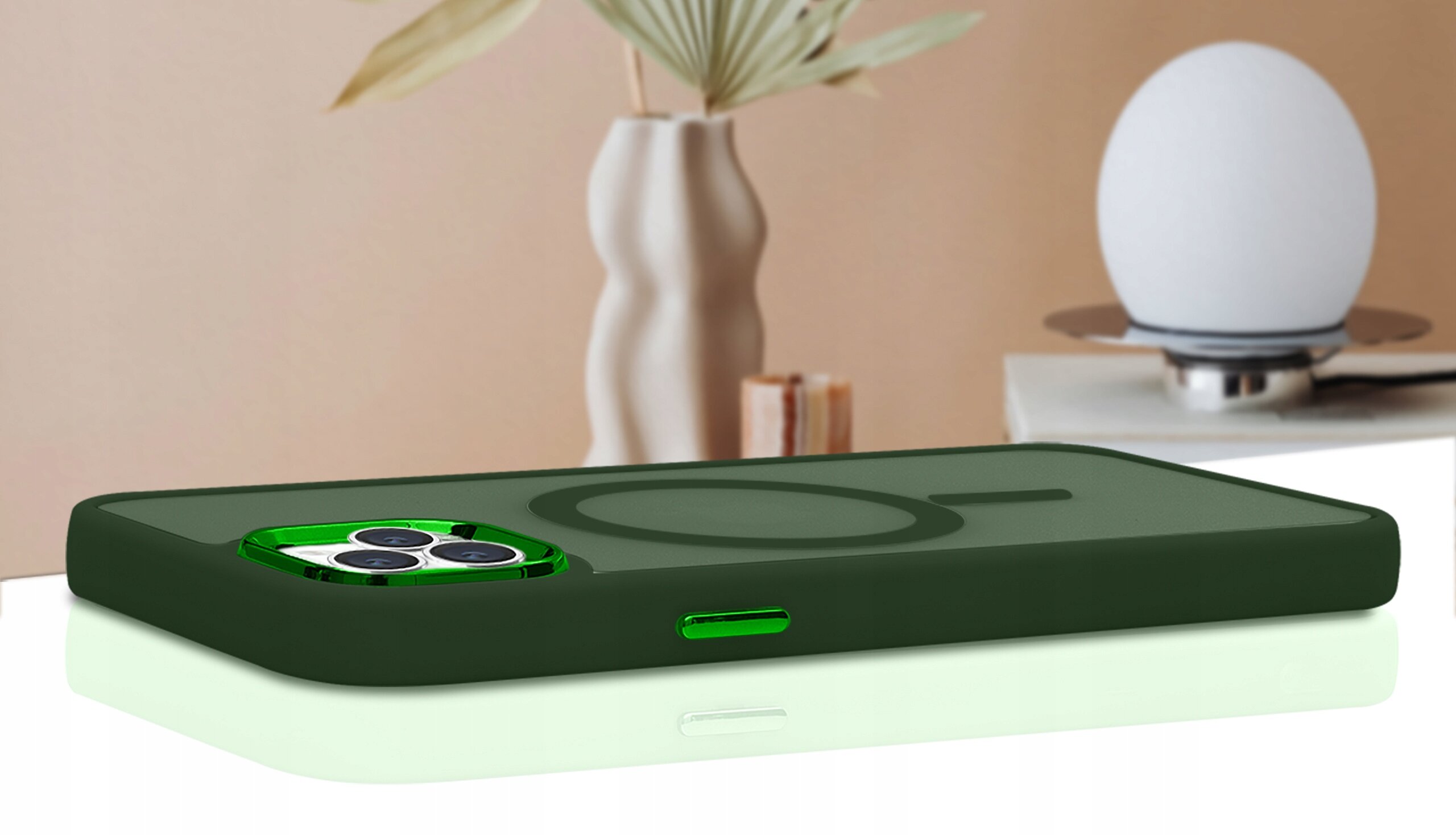 Etui do iPhone 15 Pro Max w kolorze zielonym, leży na białej powierzchni, w tle widoczny jest rozmyty element dekoracyjny w postaci wazonu i lampy.
