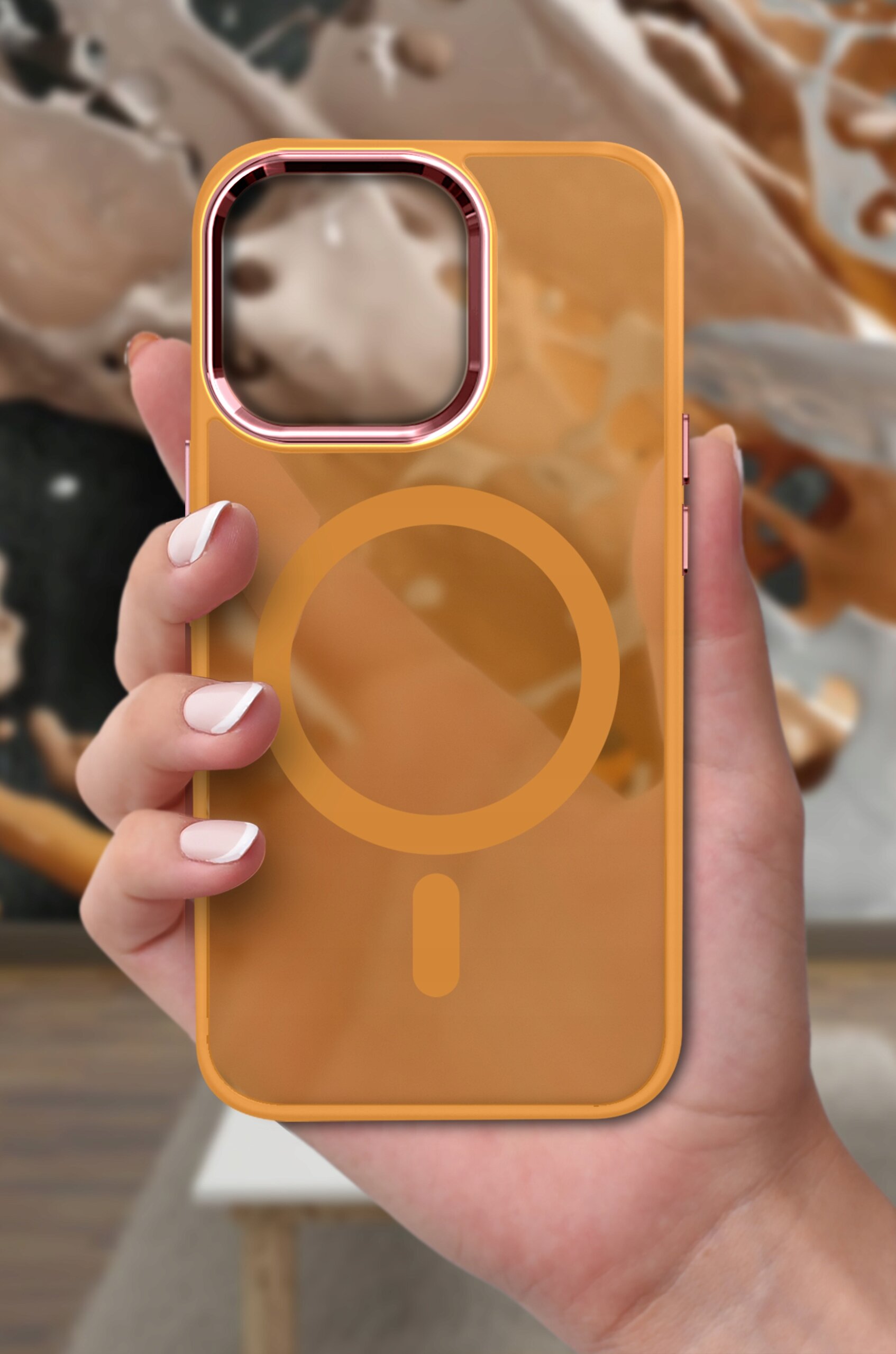 Osoba trzyma przezroczyste, pomarańczowe etui na telefon z widocznym okrągłym wzorem na środku, na tle rozmytego, abstrakcyjnego obrazu.
