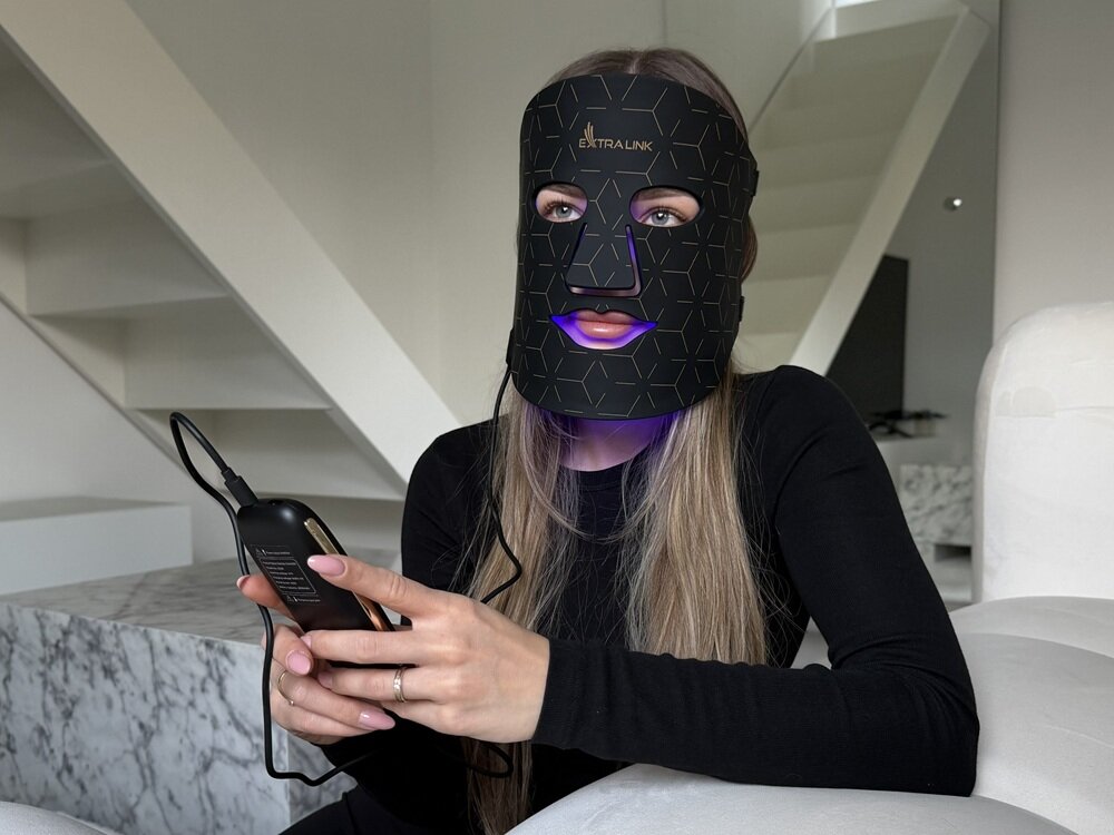Osoba siedzi na kanapie, nosząc czarną maskę LED z napisem \'EXTRALINK\' i trzymając w ręku urządzenie z przewodem. W tle widoczne są schody i fragment wnętrza mieszkania.