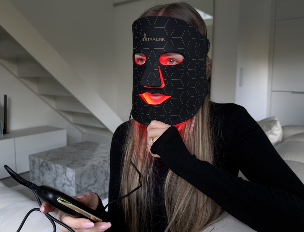 Osoba siedzi na kanapie, nosząc czarną maskę LED z czerwonym podświetleniem, na której widnieje logo \'EXTRALINK\'. W ręku trzyma urządzenie z przewodem, prawdopodobnie do sterowania maską.