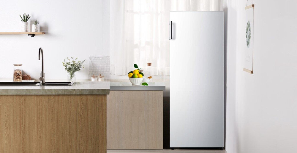 Wnętrze nowoczesnej kuchni z widoczną lodówką w kolorze inox, obok której stoi blat z umywalką i dekoracjami. Na blacie znajduje się misa z owocami, a w tle widać zasłonięte okno.