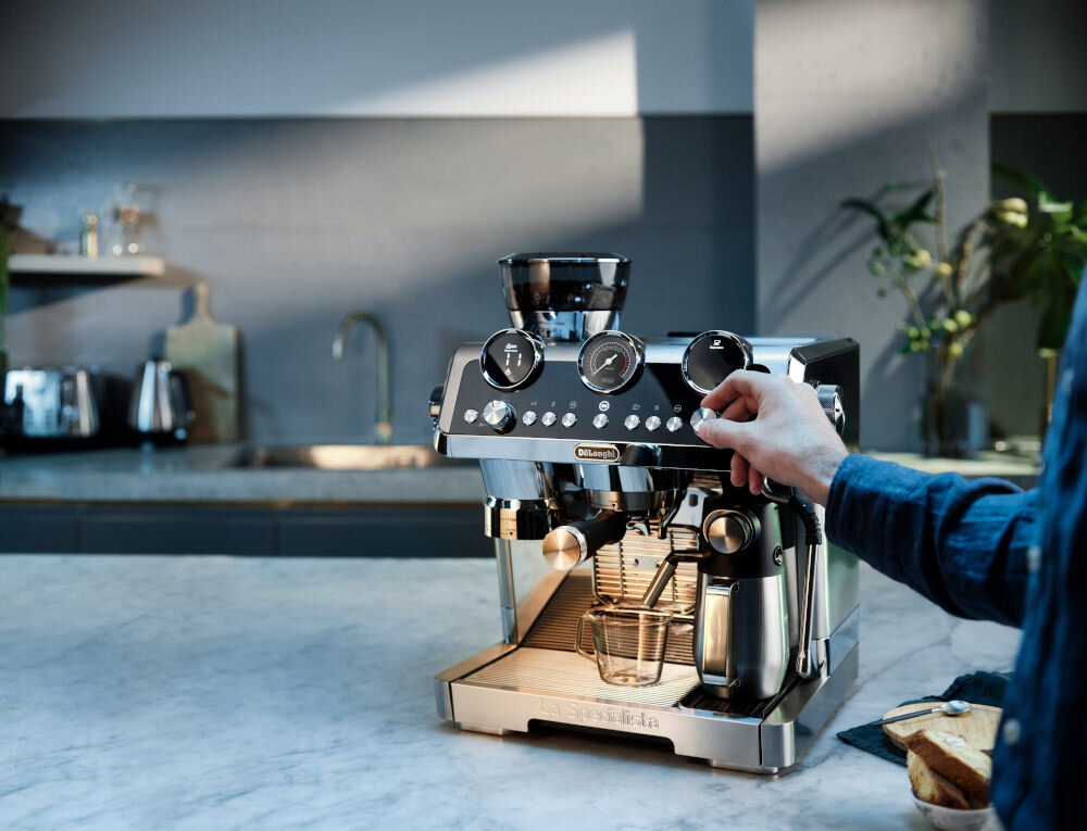 Na blacie kuchennym stoi ekspres do kawy z logo \'DeLonghi\', z widocznymi pokrętłami i wskaźnikami. Obok ekspresu znajduje się ręka osoby, która obsługuje urządzenie.