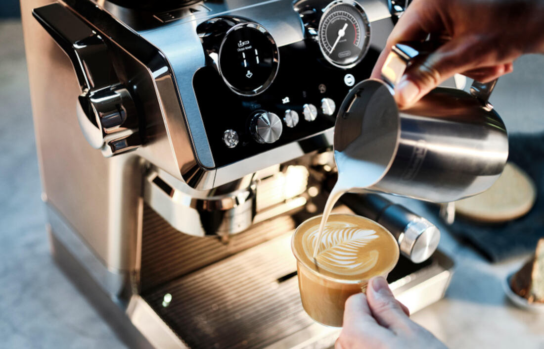 Osoba nalewa mleko z metalowego dzbanka do filiżanki z kawą, tworząc wzór latte art. W tle widoczny jest ekspres do kawy z różnymi pokrętłami i wskaźnikami.