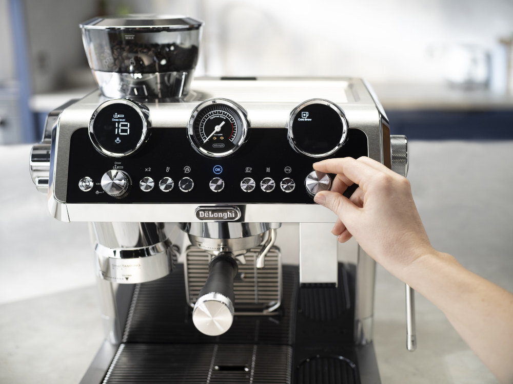 Ekspres do kawy z widocznym logo DeLonghi, posiadający pokrętła i wyświetlacze na panelu sterowania. Ręka osoby obsługującej ekspres dotyka jednego z pokręteł.