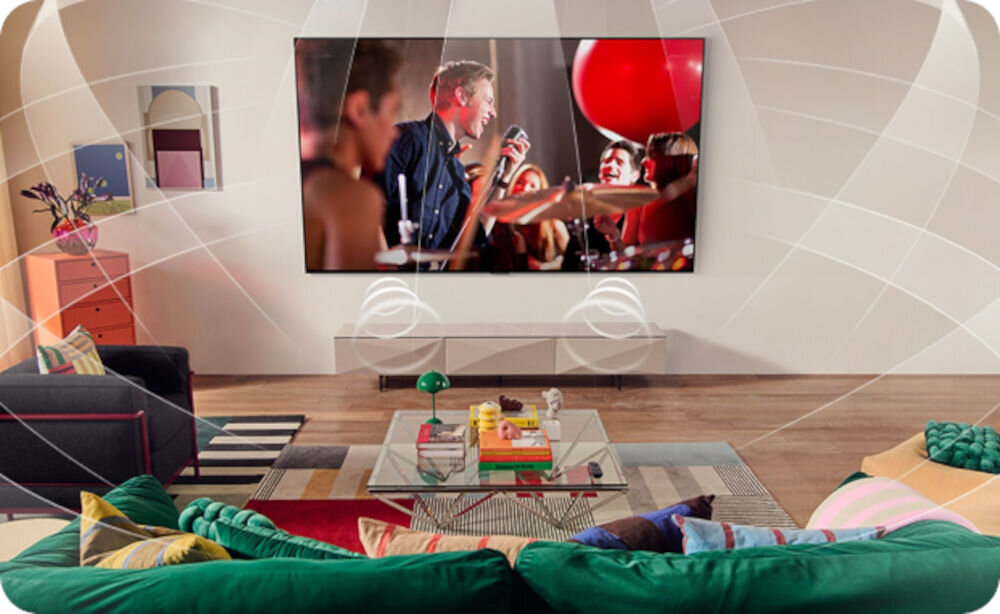 W salonie na ścianie wisi duży telewizor, na którym wyświetlany jest koncert. Przed telewizorem znajduje się kolorowa kanapa i stolik z książkami oraz dekoracjami.