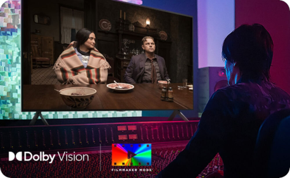 Osoba siedzi przed dużym telewizorem, na którym wyświetlana jest scena z dwoma osobami przy stole. Na dole ekranu widoczne są logotypy \'Dolby Vision\' i \'Filmmaker Mode\'.