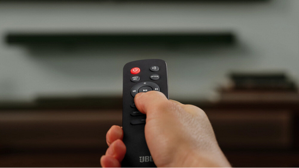 Osoba trzyma pilot z logo JBL, na którym widoczne są przyciski do sterowania urządzeniem audio.