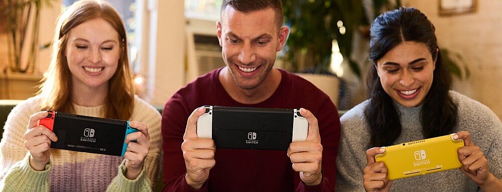 Trzy osoby trzymają konsole do gier, każda w innym kolorze: czerwono-niebieska, biała i żółta. Na konsolach widoczne jest logo Nintendo Switch.