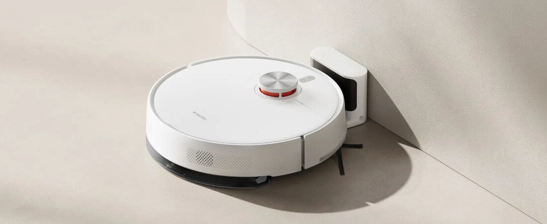 Na obrazie widoczny jest biały robot sprzątający z okrągłym kształtem, stojący przy stacji dokującej. Na powierzchni robota znajduje się logo \'Xiaomi\'.