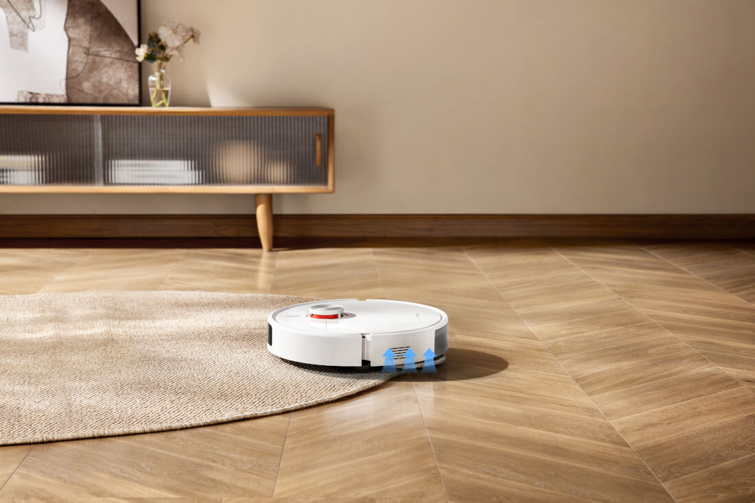 Na drewnianej podłodze znajduje się biały robot sprzątający, częściowo na dywanie. W tle widoczna jest szafka z dekoracjami.
