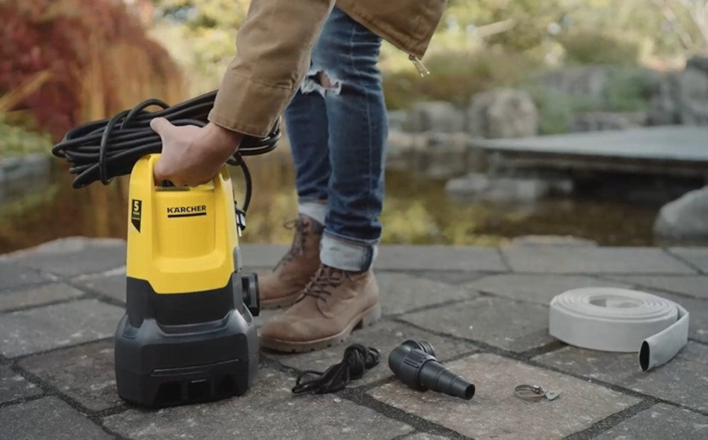 Osoba trzyma żółto-czarną pompę zanurzeniową z widocznym logo KARCHER. Obok na ziemi leżą akcesoria, w tym wąż i złączki.
