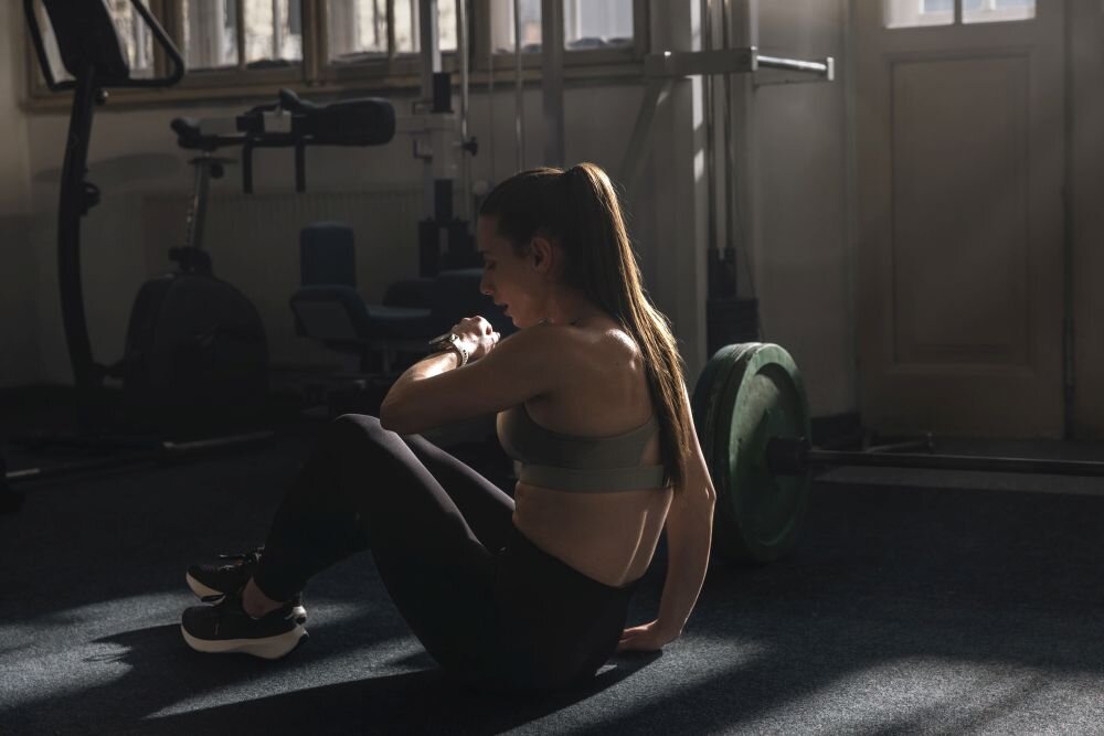 Osoba w stroju sportowym siedzi na podłodze w siłowni, patrząc na zegarek na nadgarstku. W tle widoczne są sprzęty do ćwiczeń i sztanga z obciążeniem.
