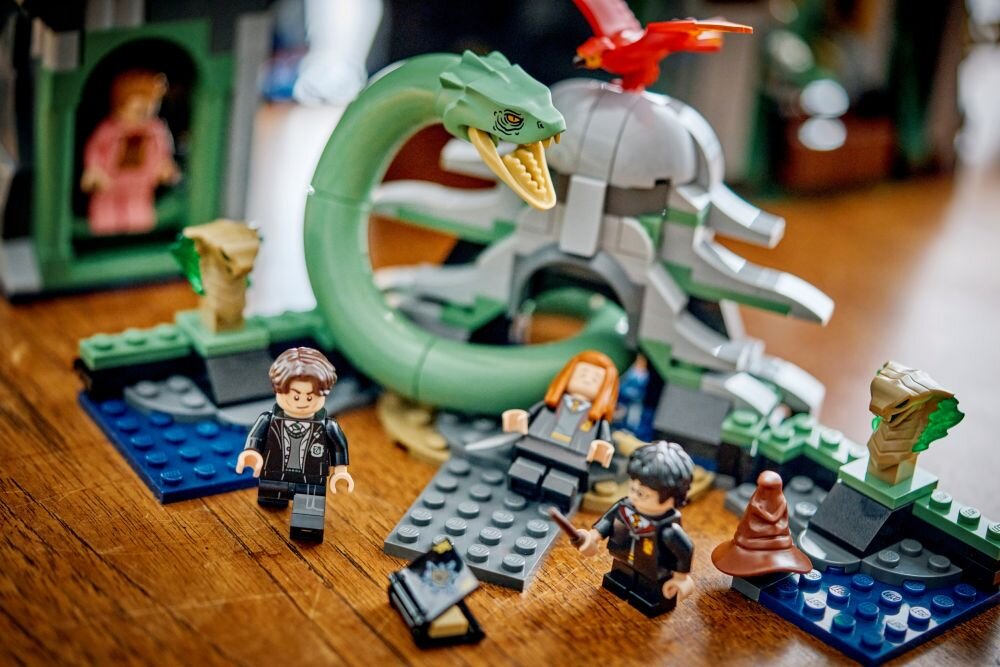 Цей набір LEGO зображує сцену з великою зеленою змією, трьома фігурками та різними конструкторськими елементами. На передньому плані видно коричневий капелюх і маленький чорний предмет.