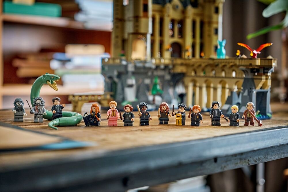 Фігурки LEGO, що зображують персонажів із серії про Гаррі Поттера, виставлені на дерев'яному столі. На задньому плані видно споруду, схожу на замок, з архітектурними деталями.