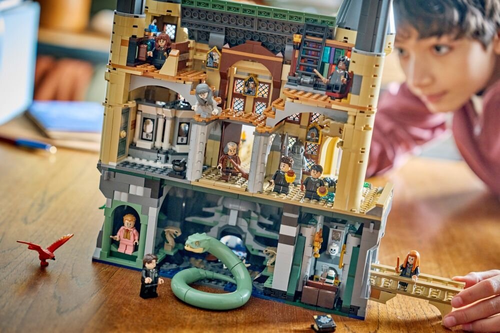 Набір LEGO із зображенням замку з мініфігурками та великою змією на дерев'яному столі. На задньому плані видно людину, яка спостерігає за будівництвом.