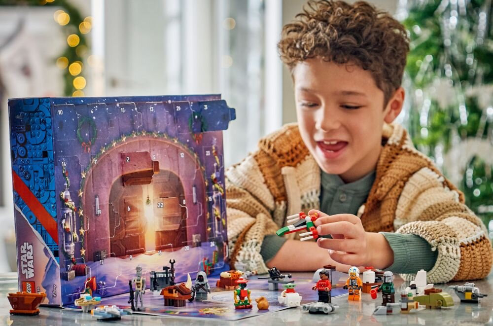Na obrazie widać dziecko bawiące się zestawem klocków LEGO, obok znajduje się kalendarz adwentowy z motywem Star Wars. W tle widoczne są świąteczne dekoracje i choinka.