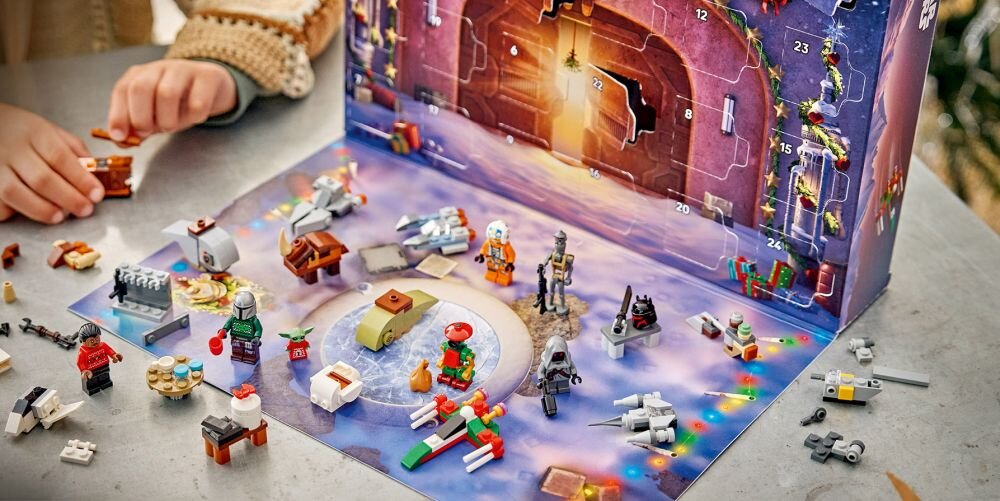 Na obrazie widoczny jest zestaw klocków LEGO z motywem Star Wars, rozłożony na stole. W tle znajduje się otwarty kalendarz adwentowy z numerowanymi okienkami.