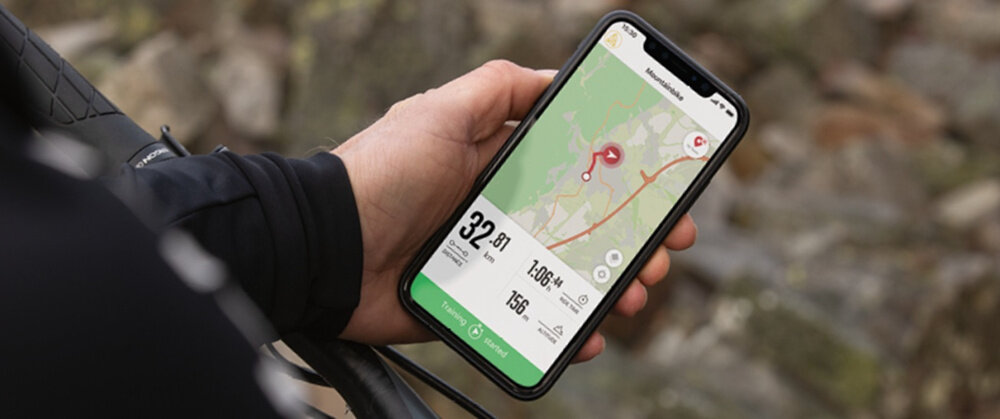 Osoba trzyma smartfon z wyświetloną aplikacją pokazującą mapę i dane dotyczące jazdy na rowerze, takie jak prędkość 32,81 km/h, dystans 1,006 km i tętno 156 bpm. W tle widoczne są rozmyte kamienie.