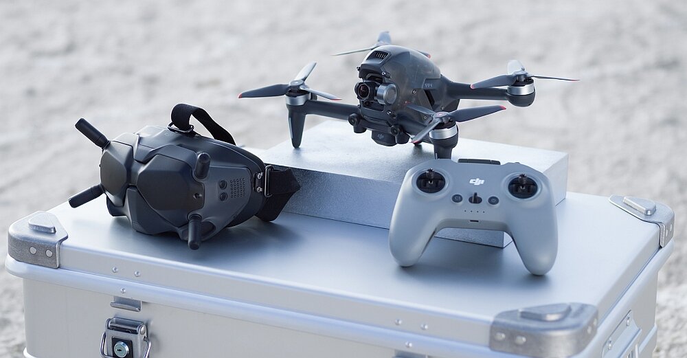 Na obrazie znajduje się dron, gogle i kontroler umieszczone na metalowej skrzyni. Dron ma cztery śmigła, a kontroler posiada logo DJI.