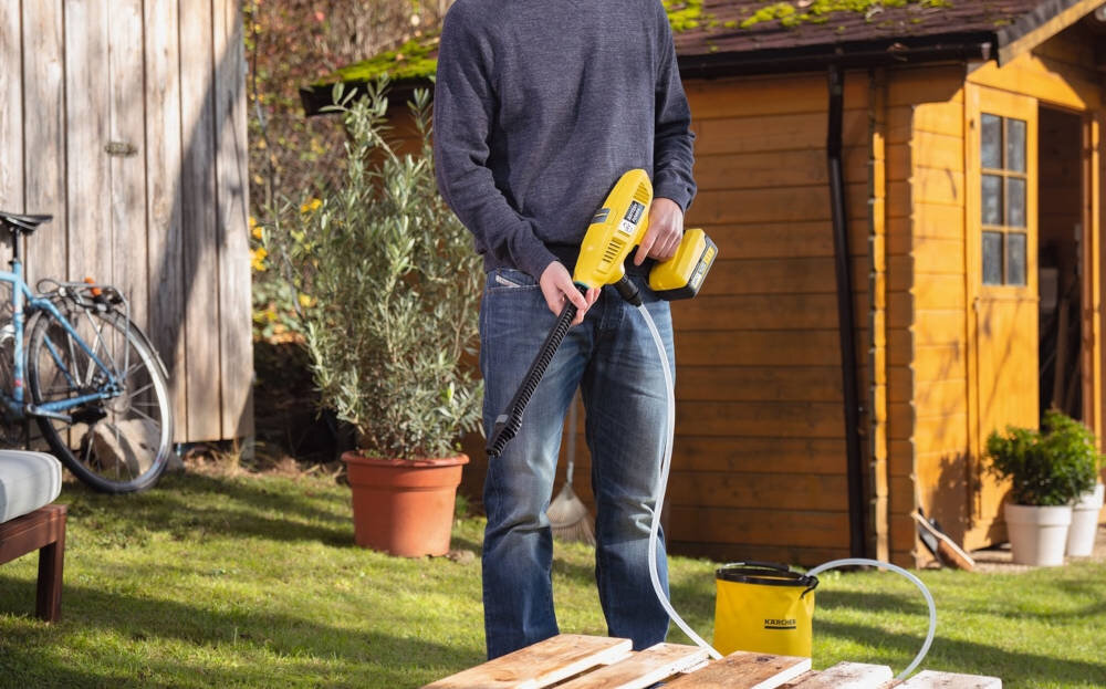 Osoba trzymająca żółtą myjkę ciśnieniową z widocznym logo KARCHER, stojąca na trawie obok drewnianego budynku. W tle widoczny jest rower i rośliny w doniczkach.