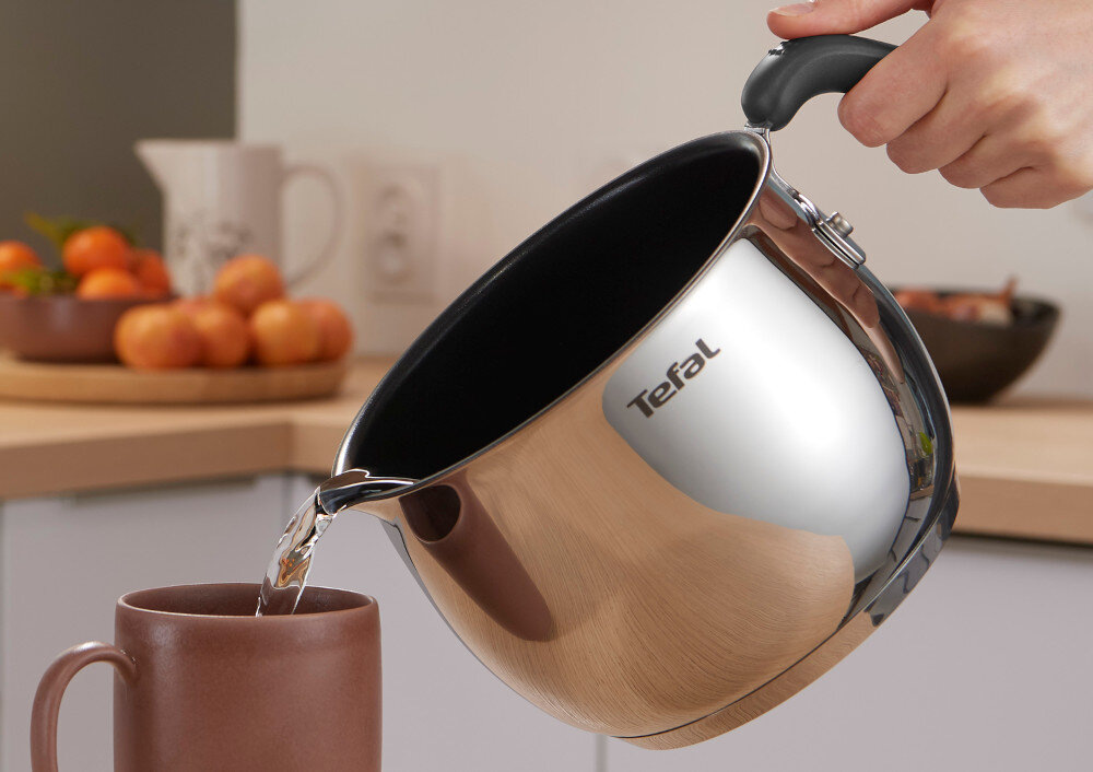 Osoba nalewa wodę z metalowego garnka z logo \'Tefal\' do brązowego kubka. W tle widać kuchenny blat z owocami i naczyniami.