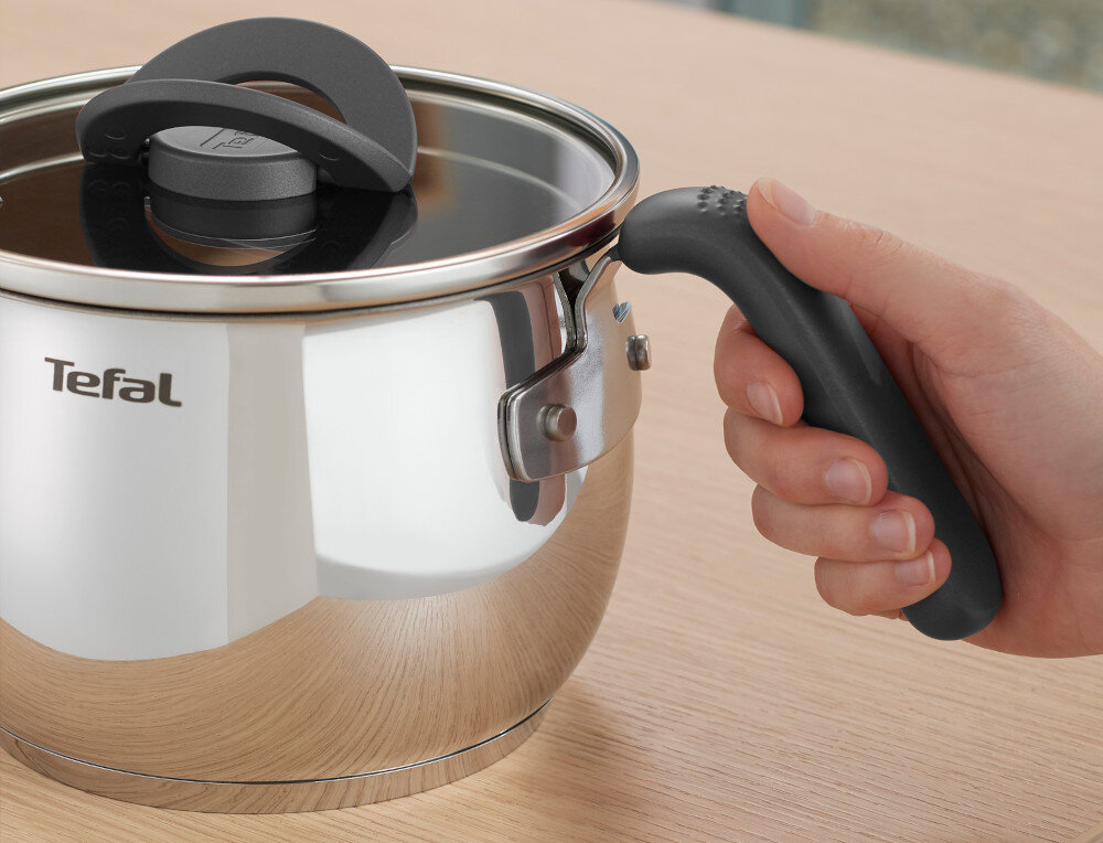 Na obrazie widoczny jest metalowy garnek z czarną pokrywką i uchwytem, trzymany przez rękę. Na garnku znajduje się logo \'Tefal\'.