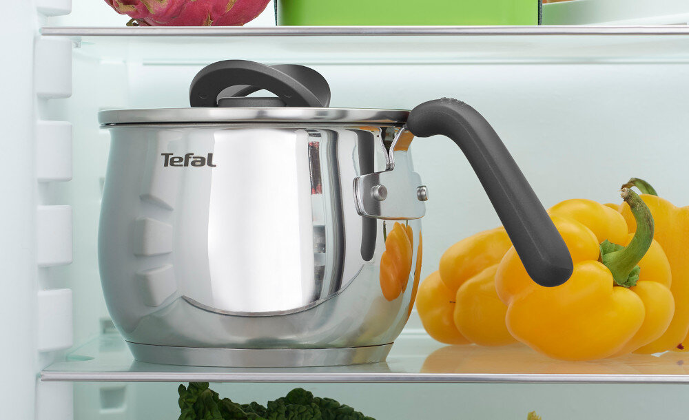 Na półce w lodówce znajduje się metalowy garnek z czarną rączką i pokrywką, obok leży żółta papryka. Na garnku widoczne jest logo \'Tefal\'.